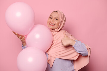 Fototapeta na wymiar Stylish elegant middle-aged pretty Muslim woman in a hijab, gesturing with a thumb up, smiling, posing with air balloons, expressing happiness and positive emotions on isolated pink color background