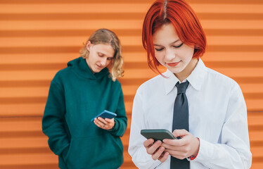 Smiling caucasian teenage girl portrait with red dyed hair and friend boy behind browsing their smartphone devices. Careless young teenhood time and a modern technology concept image.