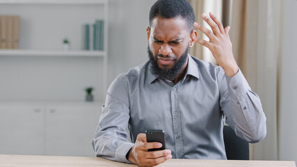 Stressed mad angry African American ethnic bearded man with mobile phone reading bad news. Sad...