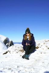 Fototapeta na wymiar A young attractive woman sits on the snow, holds a cup with a drink in her hands and smiles. Full length portrait of a female tourist on winter vacation