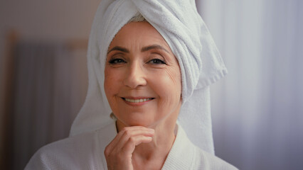 Cheerful female portrait happy 50s middle-aged lady 60s mature woman touching facial skin looking at camera with smile touch face pampering wears towel on head enjoy cosmetics and plastic surgery