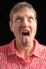 Paris, France - 03 04 2022: Studio shot of a mature woman grimaces sticking her tongue out and looking up, with short hair wearing a pink and white shirt with cufflinks