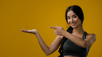 Portrait in studio Indian 20s woman posing on yellow background pointing to side at palm arm hand point aside recommend recommendation sign girl lady showing to empty copy space looking at camera