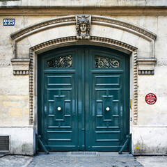 Paris ancient stone building entrance with wooden carved double doors, brass handles, stone walls, rich stucco fretwork, street number, ventilation grille, red traffic sign and cobblestone pavement