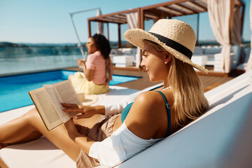 Young woman reads book while relaxing by pool on summer vacation. - Powered by Adobe