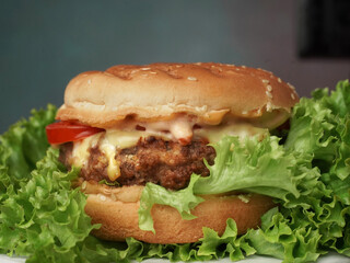 Close up of Big Burger lies on craft white paper against wooden table. A Juicy green Salad leaf and a red Tomato lie near the Burger