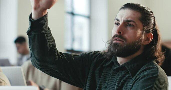 Close up of handsome Caucasian young man with beard sitting at lection in University, rising hand and asking question or anwering to professor. Communiction between student and teacher.