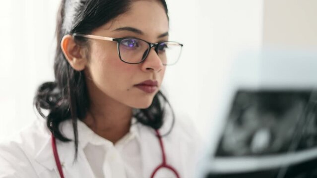 Portrait of concentrated indian woman doctor therapist with glasses thinking about diagnosis while looking at results patient MRI or CT scan procedure at light hospital office