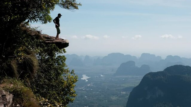 Tripod shot of a tourist standing and sitting on the cliff edge overhanging the plain on sunny day. Dragon's Crest (Khao Ngon Nak) Viewpoint, Krabi Province, Thailand.