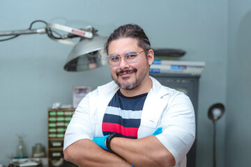 A local practicing doctor running a small animal bite center or small clinic. Posing after a long day, wearing a short sleeved smock, with a casual shirt underneath and gloves.