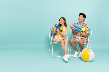 Young Asian couple in summer outfits sitting on a beach chair and holding water guns plastic for...