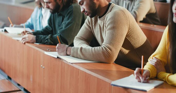 Close up of mixed-races males and females students sitting at desk in class and writing down information with professor explanation. Inside. Multiethnic people noting at lesson. College lifestyle.