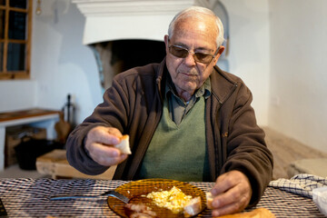 retired elderly man eating fried egg at table with chequered tablecloth, old earthen fireplace in...