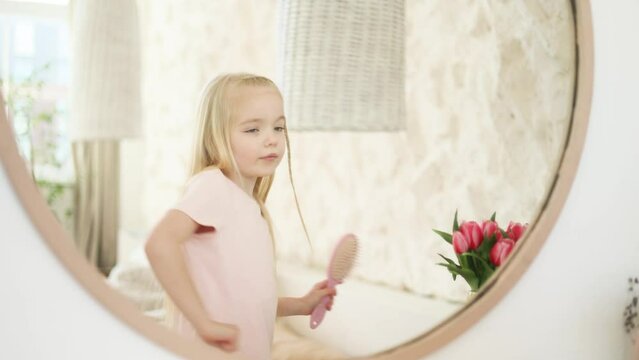 Adorable funny expressive little girl child with hair brush looking at mirror reflection dancing and singing at home Cute blond kid enjoying free time in light room in morning before school indoors