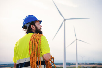 Back view of windmill turbine maintenance engineer standing wearing a hard hat nd a safety vest at...