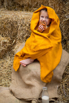 A little girl with bare feet, wrapped in an orange linen, sits on a burlap on a stack of straw and eats a cinnamon bun