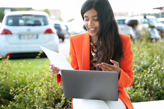 Young smiling Indian business woman using laptop computer holding contract having video call on street. Confident asian freelancer wearing stylish jacket working online project sitting at workplace