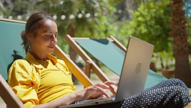 Side view remote online working woman sitting on sun lounger with palm trees on background, traveler blogger using laptop computer, connecting to internet on netbook