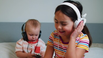 Two cheerful children in headphones sing songs and listen to music at home. Two cute children baby boy and girl seven years old spend the weekend at home listening to music in headphones.  - Powered by Adobe