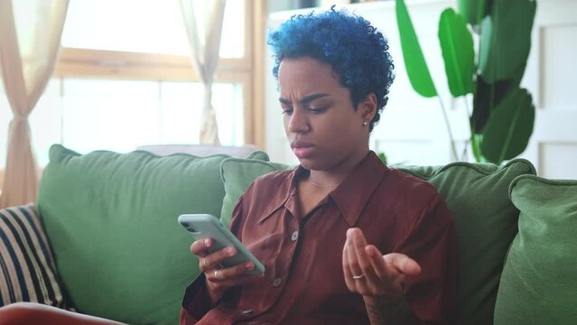 Young annoyed African American woman after reading negative news on phone hrows up hands indignantly learning about cancellation of concert or entertainment festival sits on couch in appartment