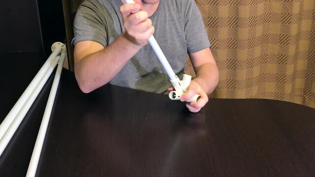 A man demonstrates parts for a garden awning. Connects tubes and adapters. Medium plan..
