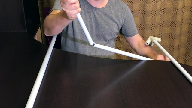 A man demonstrates parts for a garden awning. Connects tubes and adapters. Medium plan