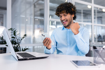 Young successful businessman inside the office at work uses a laptop hispanic man holds a smartphone in his hands, the man holds his hand up a gesture of success and triumph, celebrating victory.