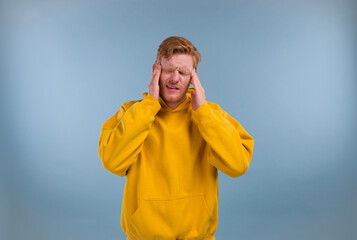 Young man wearing yellow hoodie over blue background with hand on head for pain in head because stress. suffering migraine.