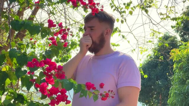 Portrait of handsome young allergic man is suffering from pollen allergy or cold on natural flowers, flowering tree background at spring or sunny summer day, sneezes, blowing his runny nose rubs eyes
