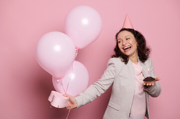 Charming birthday girl, a middle-aged multi-ethnic elegant happy woman with a festive hat on her head, holding a chocolate cake with candle and pink balloons, expressing happiness on pink background