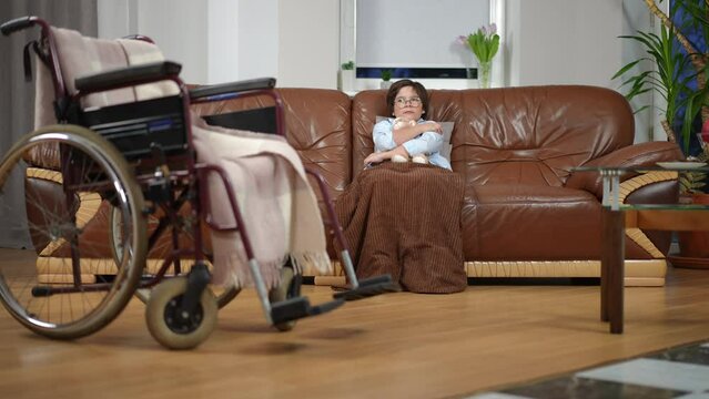 Wide shot sad ill boy hugging toy sitting on couch with blurred wheelchair at front in living room. Portrait of frustrated desperate Caucasian kid at home indoors