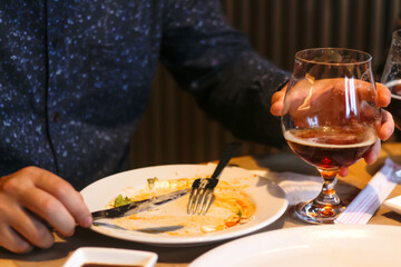 Fototapeta na wymiar After meal concept. Dirty dishes from the tables in restaurant. Empty dirty plate after meal with fork inside next to napkins with chopsticks, male hand. Man holding glass with beer or whiskey