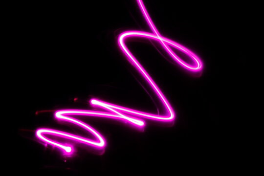Lìneas de color rosa con luz de neòn en movimiento de càmara y bokeh, forma  un garabato, formando una  ilustraciòn abstracta en el espacio con fondo negro.