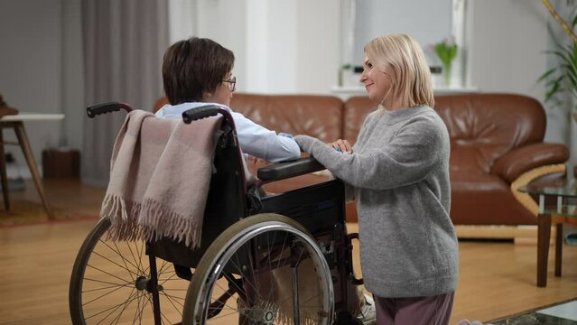 Loving mother holding hand of son in wheelchair talking smiling. Portrait of Caucasian woman taking care of ill son at home indoors. Parenting and disability concept