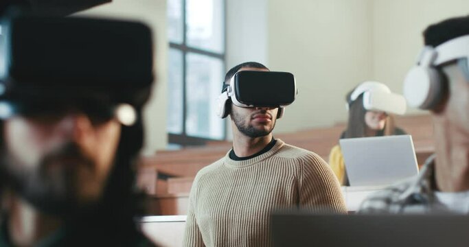 Multiethnic males and female students in VR glasses listening to lection at University and turning heads left and right. Hi-tech education. Mixed-races young people learning with virtual reality.
