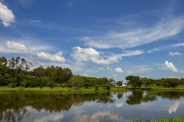 Fototapeta na wymiar Uma paisagem do Parque Leolídio di Ramos Caiado na cidade de Goiânia com um pequeno lago e o céu refletido na agua.