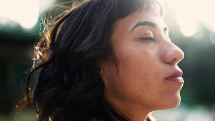 A hispanic latin woman taking a deep breath standing outside in sunlight opening eyes to sky feeling freedom
