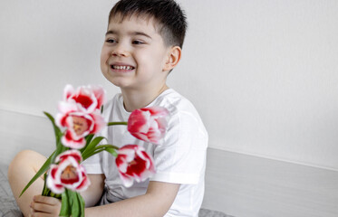 cute kid with bouquet of red tulips gift for mother women's day.child holding 2 tulips on eyes smiling having fun smelling one flower sitting on bed.birthday flowers easter spring coming preschooler 