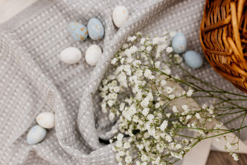 Obraz na płótnie Canvas A wicker brown basket on a gray linen napkin, are painted blue and white eggs and white small flowers. Spring background for Easter. Mock up. Space for text