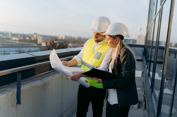 an architect and an engineer are looking at a project on the roof of a built office. male and...