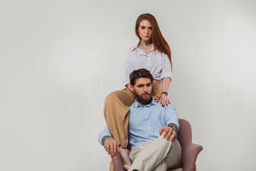 Stylish beautiful young couple brutal fashion man and a beautiful red -haired girl in a shirt sits on a chair on a white background