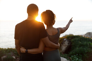 Young african american woman hugging man in sportswear, enjoy workout break, point finger at empty...
