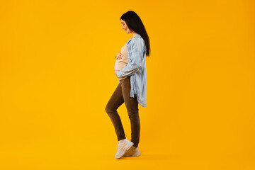 Fototapeta na wymiar Young expecting lady touching, looking at her belly and smiling, standing over yellow background, side view, full length