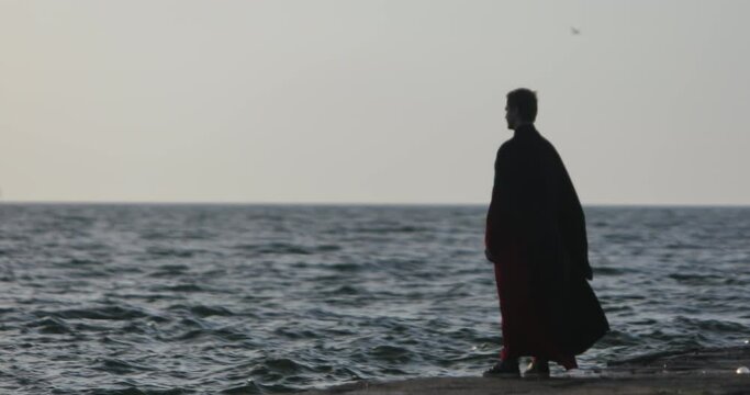 A man in a black mantle stands on a sea pier, side view