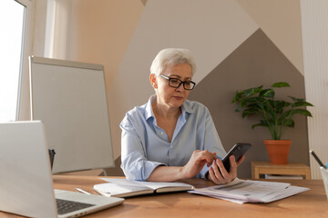 Fototapeta na wymiar Confident stylish european middle aged senior woman using smartphone at workplace. Stylish older mature 60s gray haired lady businesswoman with cell phone in office. Boss leader using internet apps.
