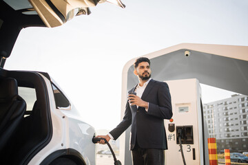 Well-dressed arabian man enjoying hot aroma coffee while charging battery of white electric car at urban station. Concept of consciousness and eco friendly auto.