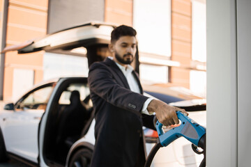 Good-looking indian man in business wear standing at charging station and taking electrical connector for charging battery of modern car. Alternative transport and ecology concept.