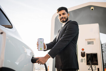 Positive arabian man in business suit charging his electric car at outdoor station and using modern...
