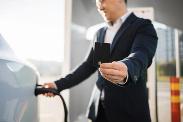 Positive asian man in stylish black suit charging white electric car and showing black credit card outdoors. Cashless payment for energy at EV station.