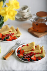 Thin pancakes with strawberries and blueberries, delicious breakfast. Homemade french crepes with fresh fruits, teapot with berries tea and bouquet of yellow flowers on table.